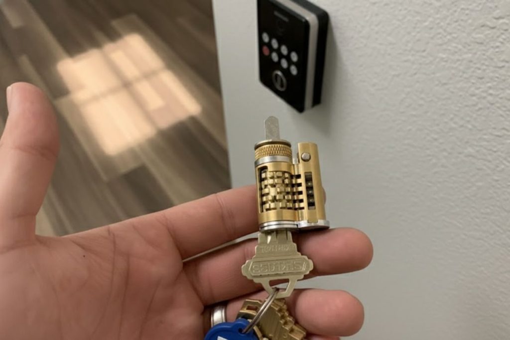 Hand holding a cutaway lock cylinder with a key inserted, showing the internal pins, with an electronic keypad door lock in the background.
