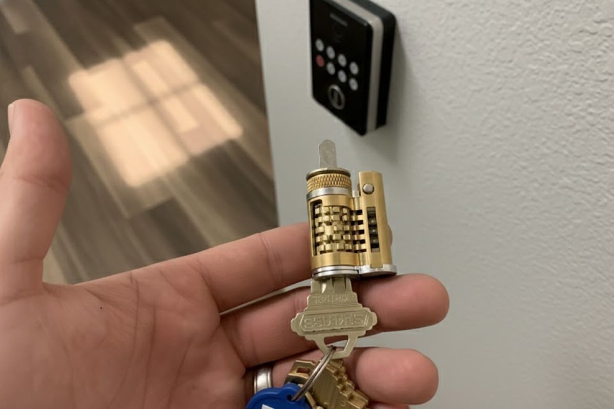 Hand holding a cutaway lock cylinder with a key inserted, showing the internal pins, with an electronic keypad door lock in the background.